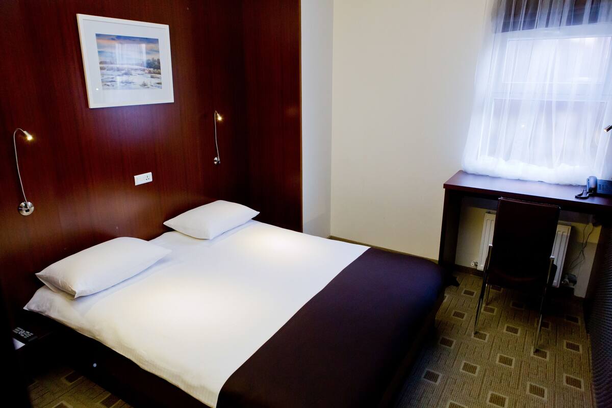 A hotel room with a bed, a desk, a chair, a picture on the wall, and a window with sheer curtains.