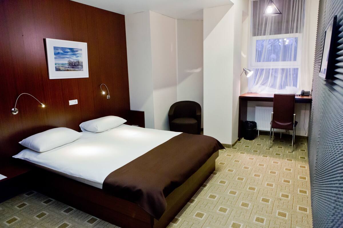 A hotel room with a bed, a desk, a chair, and a window with sheer curtains.