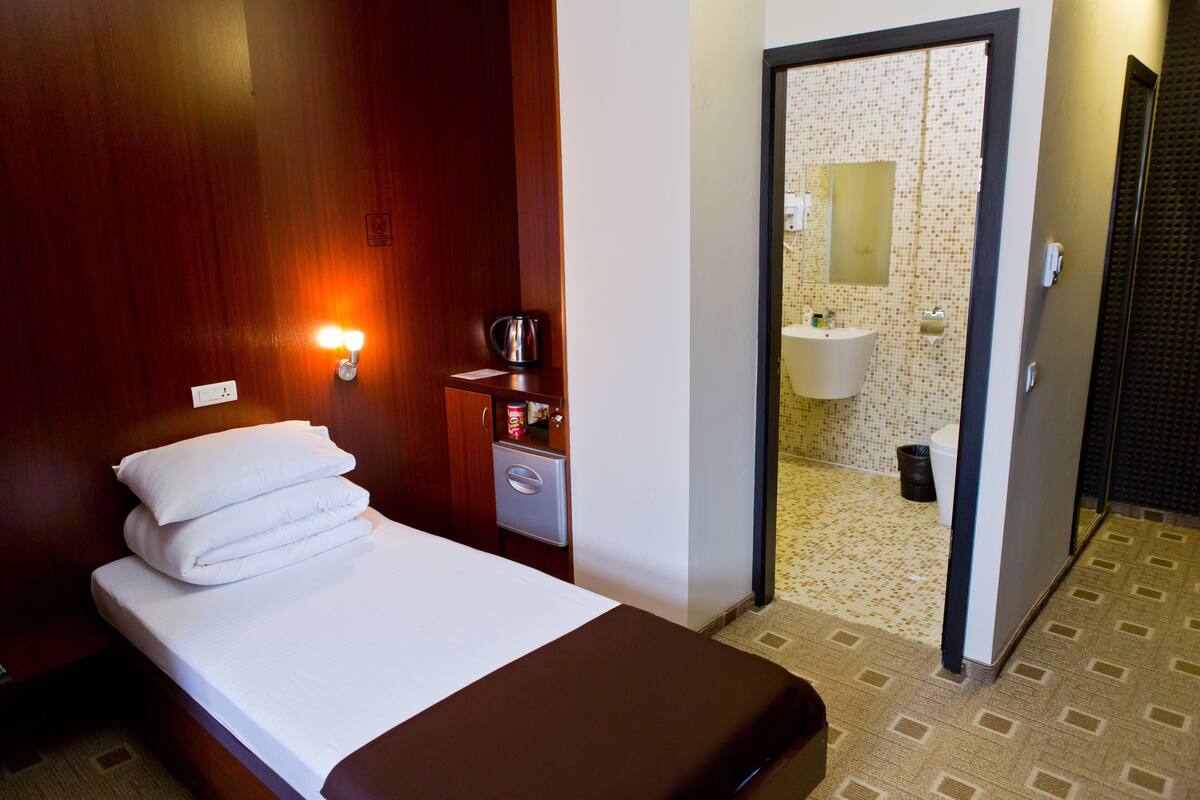 A hotel room with a bed, a nightstand with a lamp, a coffee maker, and a view of the bathroom through an open door.
