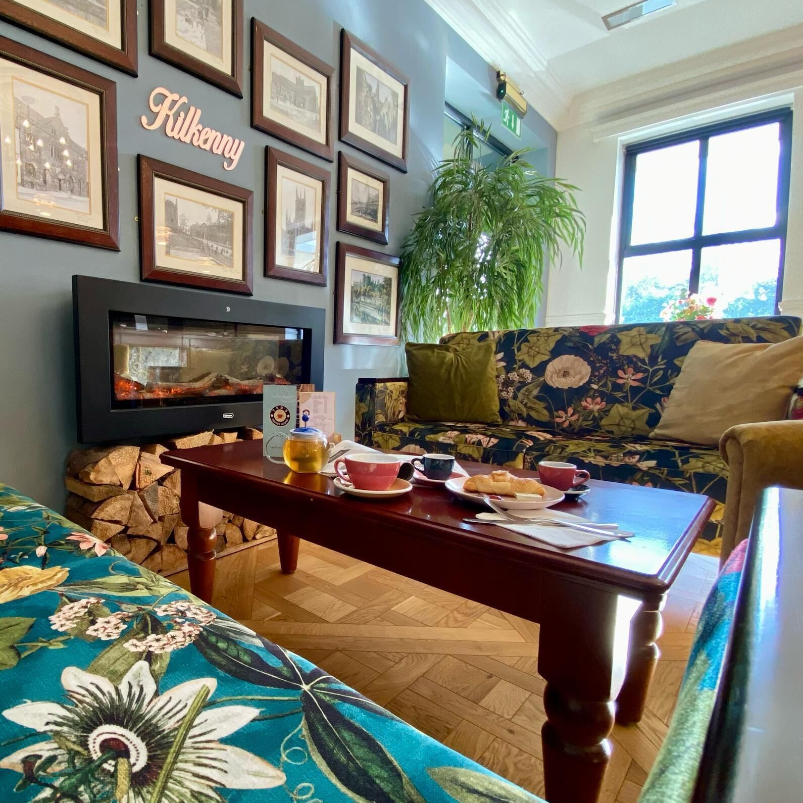 Una sala de estar acogedora con chimenea, una mesa de centro con alimentos para el desayuno, un sofá con tapicería floral y una pared decorada con cuadros.