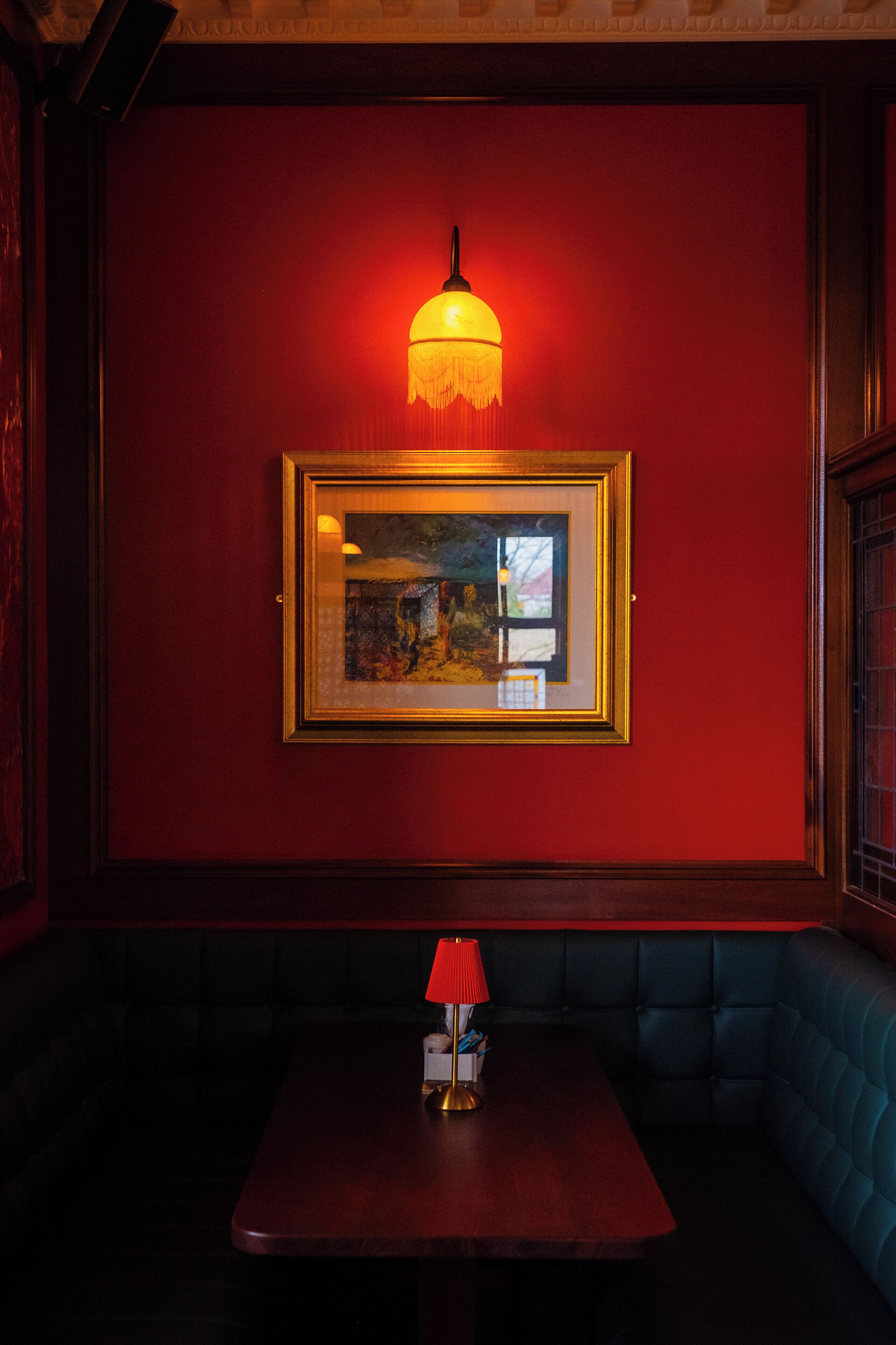 Een rode muur met een ingelijst schilderij, een tafel met een rode lamp, en een blauwe met kussens beklede bank.