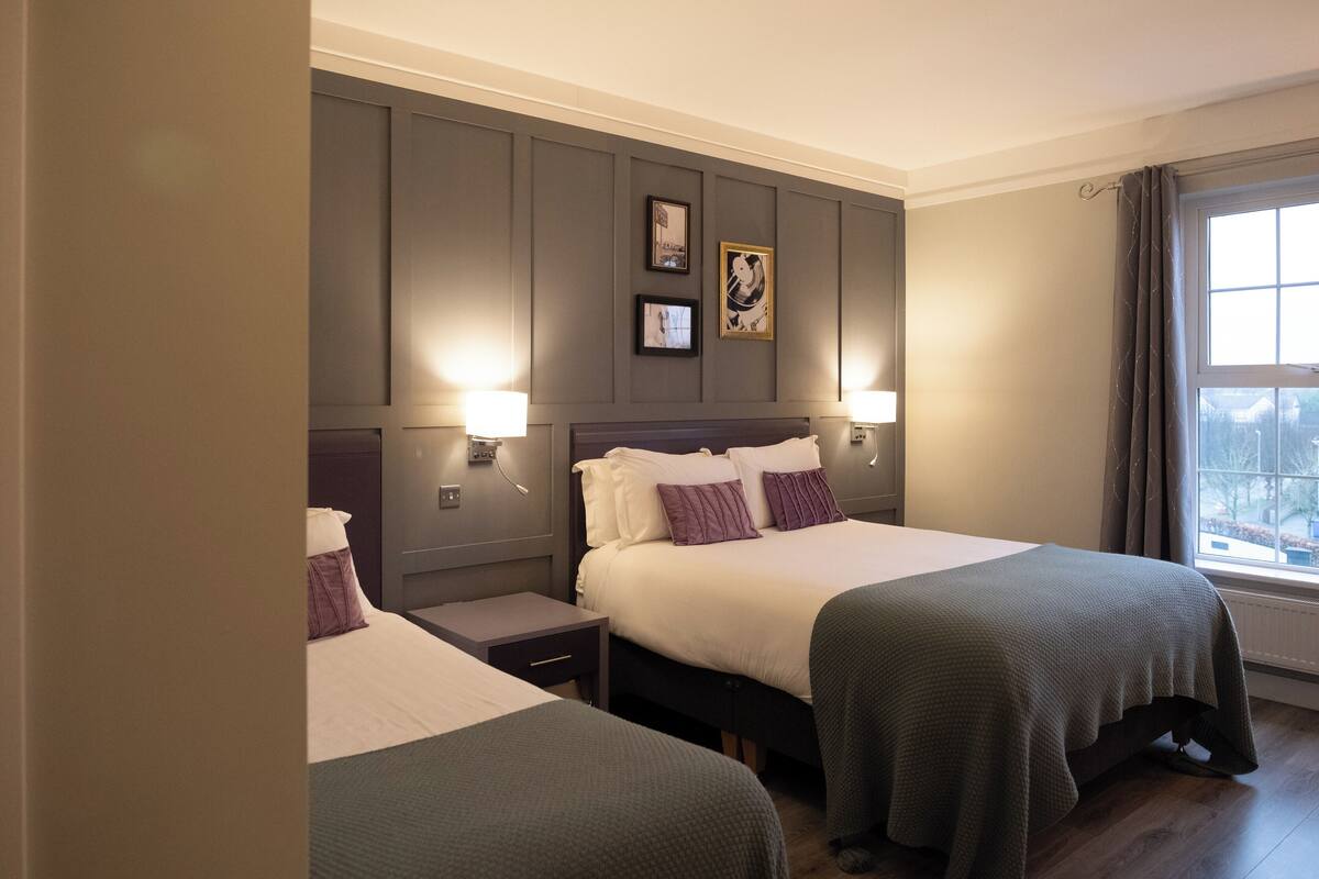 Habitación de hotel con dos camas, un ventanal grande con cortinas y lámparas fijadas a la pared.
