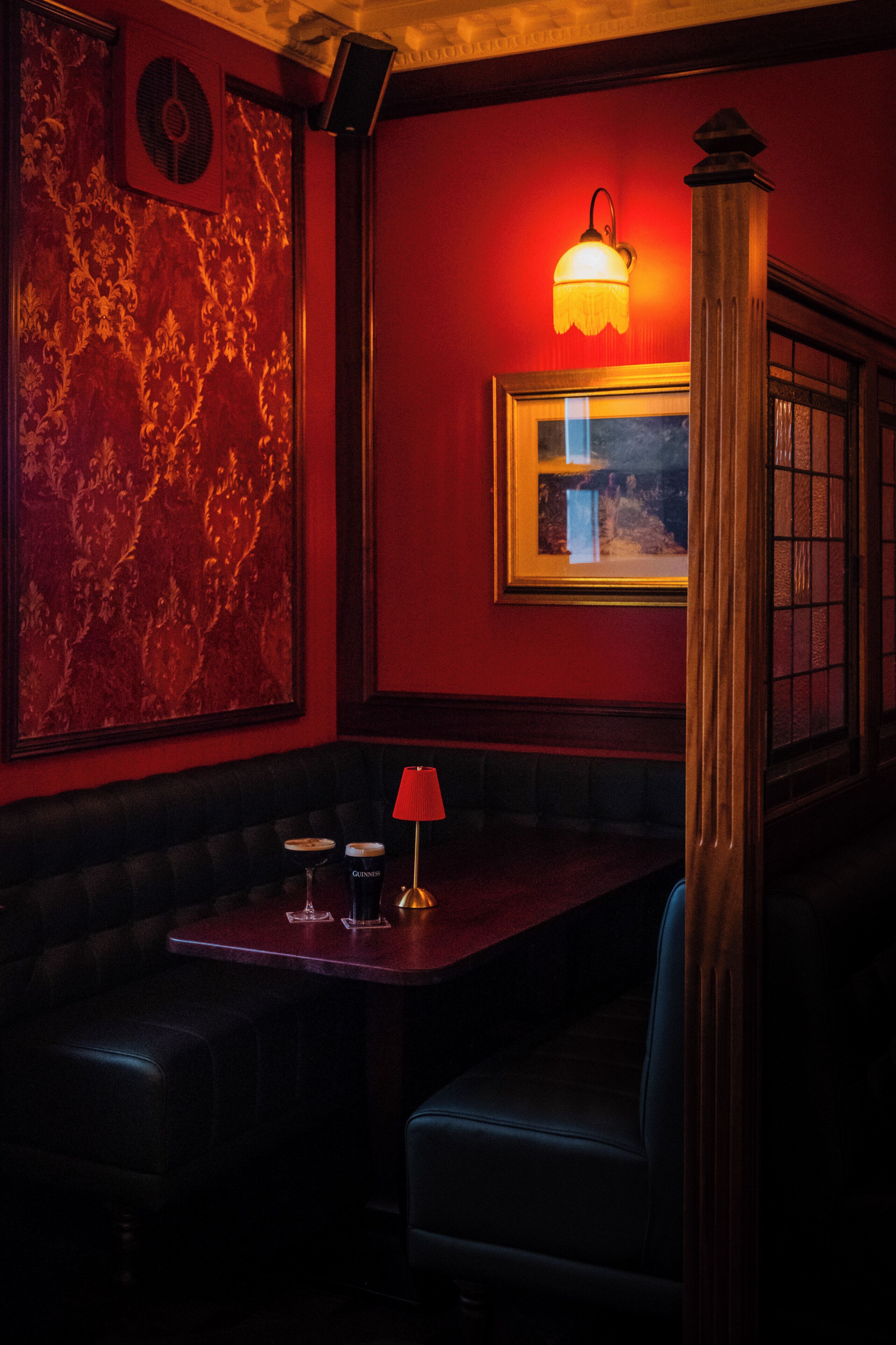 A dimly lit room with a red wall, a large framed picture, a table with a red lamp, and a window with a view.