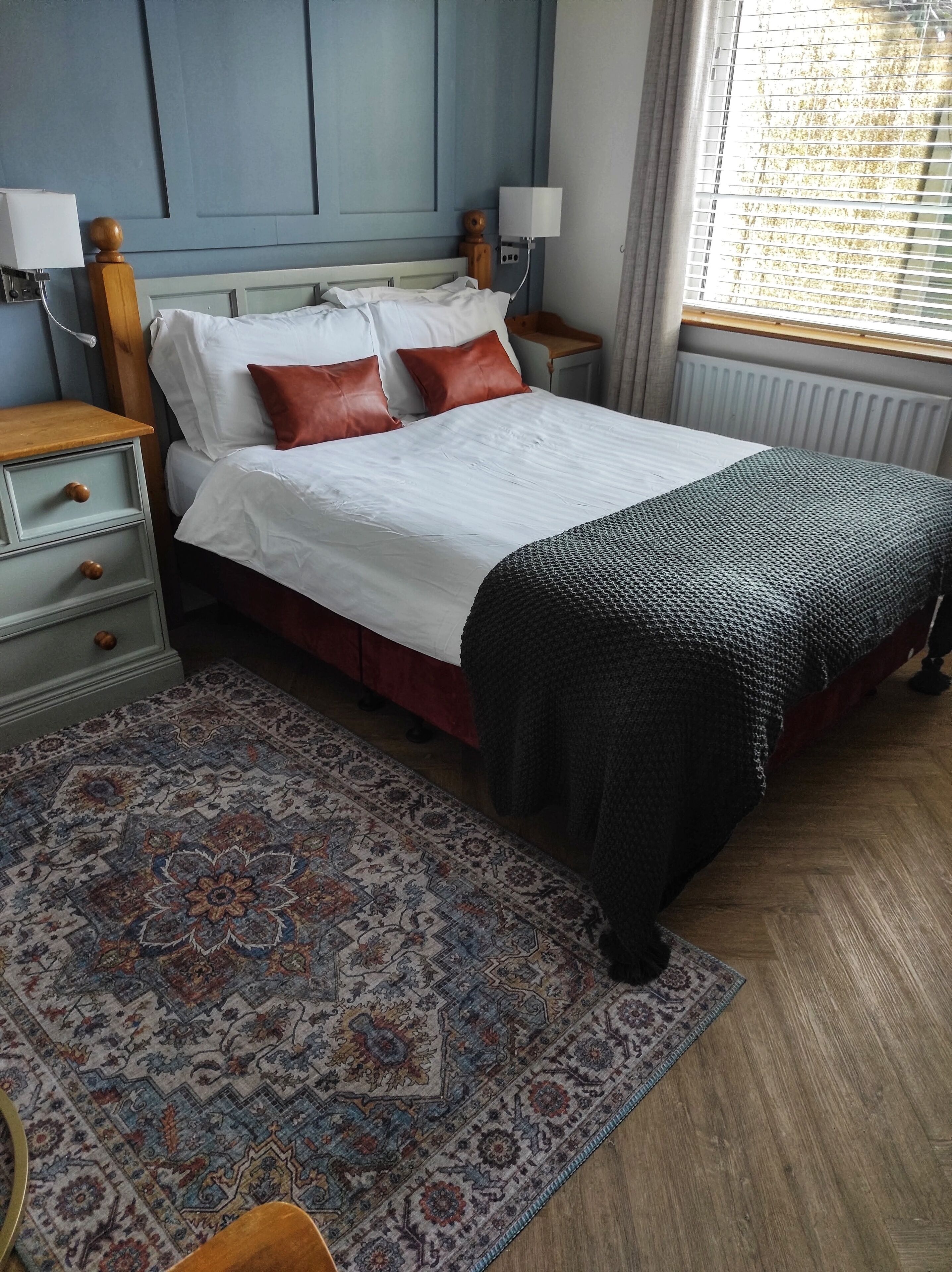 Una cama bien hecha con ropa de cama blanca y marrón, una alfombra estampada, mesitas de noche de madera y una ventana con persianas.