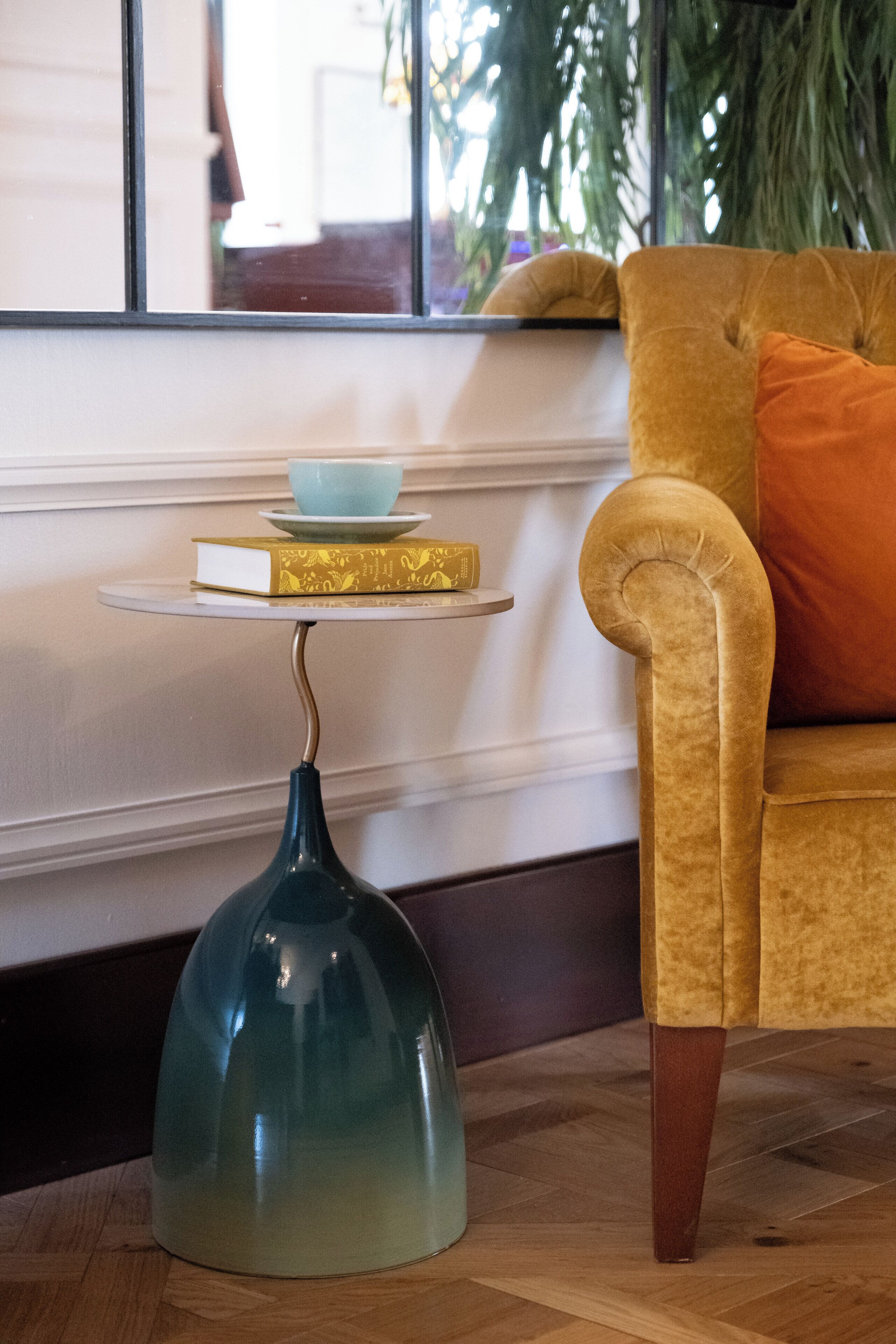 Una habitación con un sillón amarillo, una mesita con un tazón azul y un libro, y un jarrón verde.