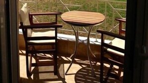 A balcony with two wooden chairs and a small round table, overlooking a residential area with houses and a road. - Pfaffenhofen (Volvi)