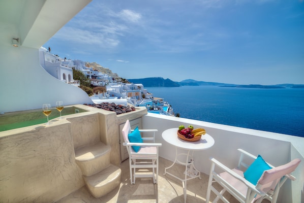 A balcony with white furniture, a table with fruit, and a view of the sea and white buildings. - Kima Oia Suites (Santorini)