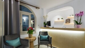 Un área de recepción con mostrador curvo, dos sillones con cojines estampados, una planta en maceta con flores rojas y vista al exterior por una ventana.