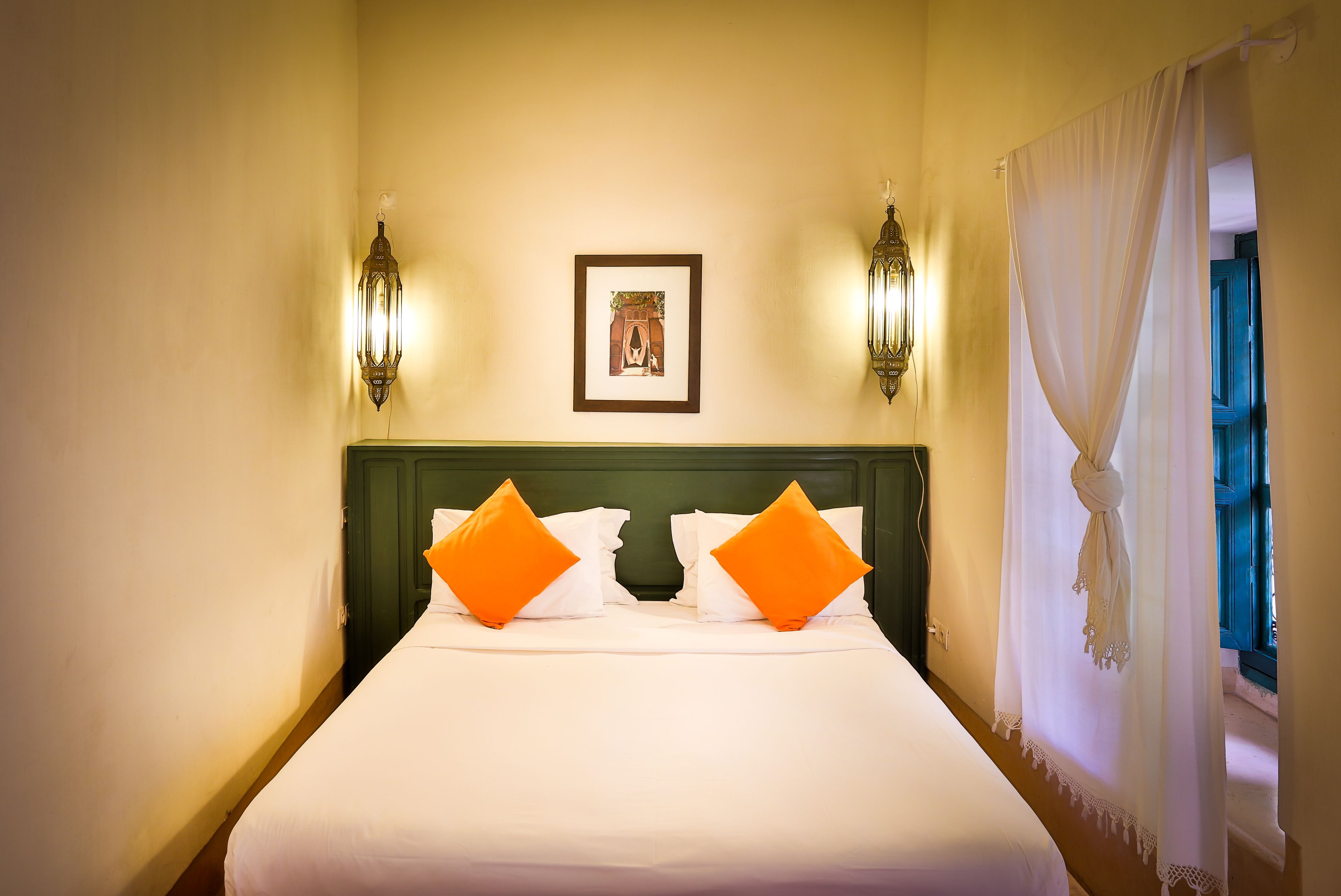 A neatly made bed with white bedding and two orange pillows, flanked by wall-mounted light fixtures and a framed picture above.