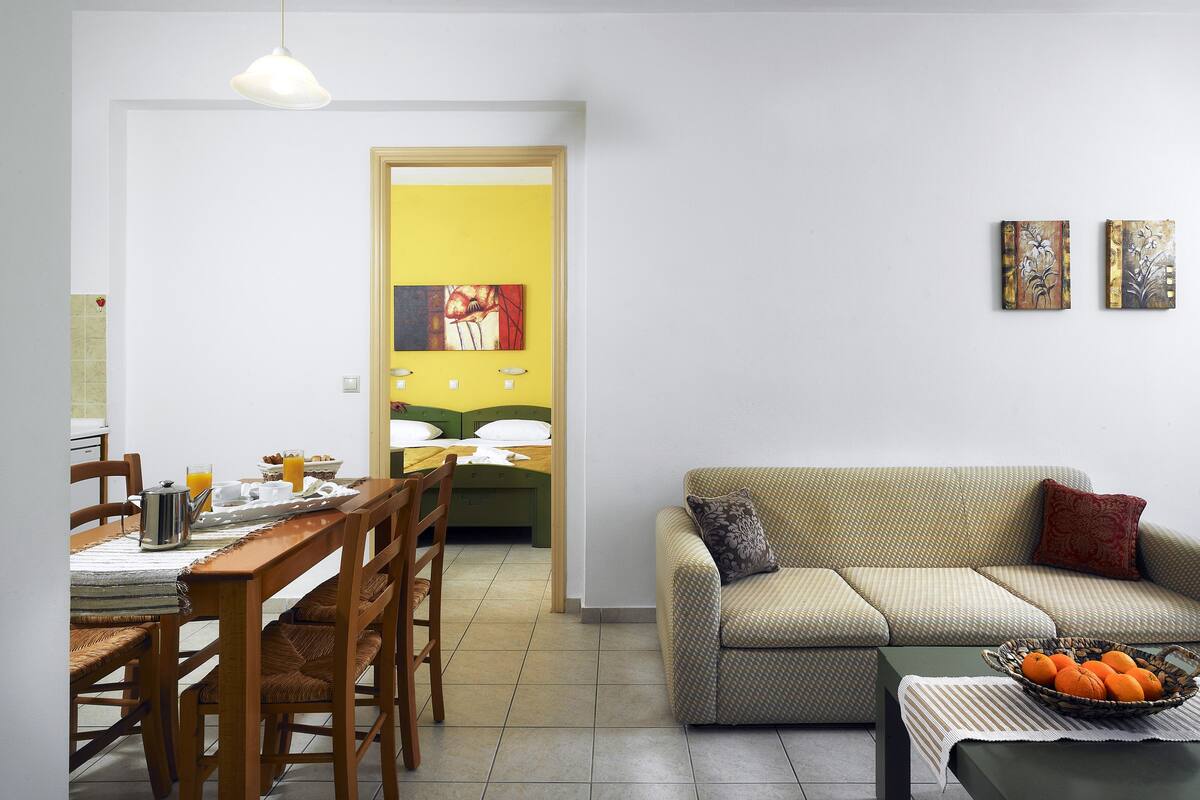Un coin repas avec une table et des chaises en bois, une cuisine visible à travers une porte ouverte, un canapé avec des coussins et un panier de fruits posé sur une table d’appoint.