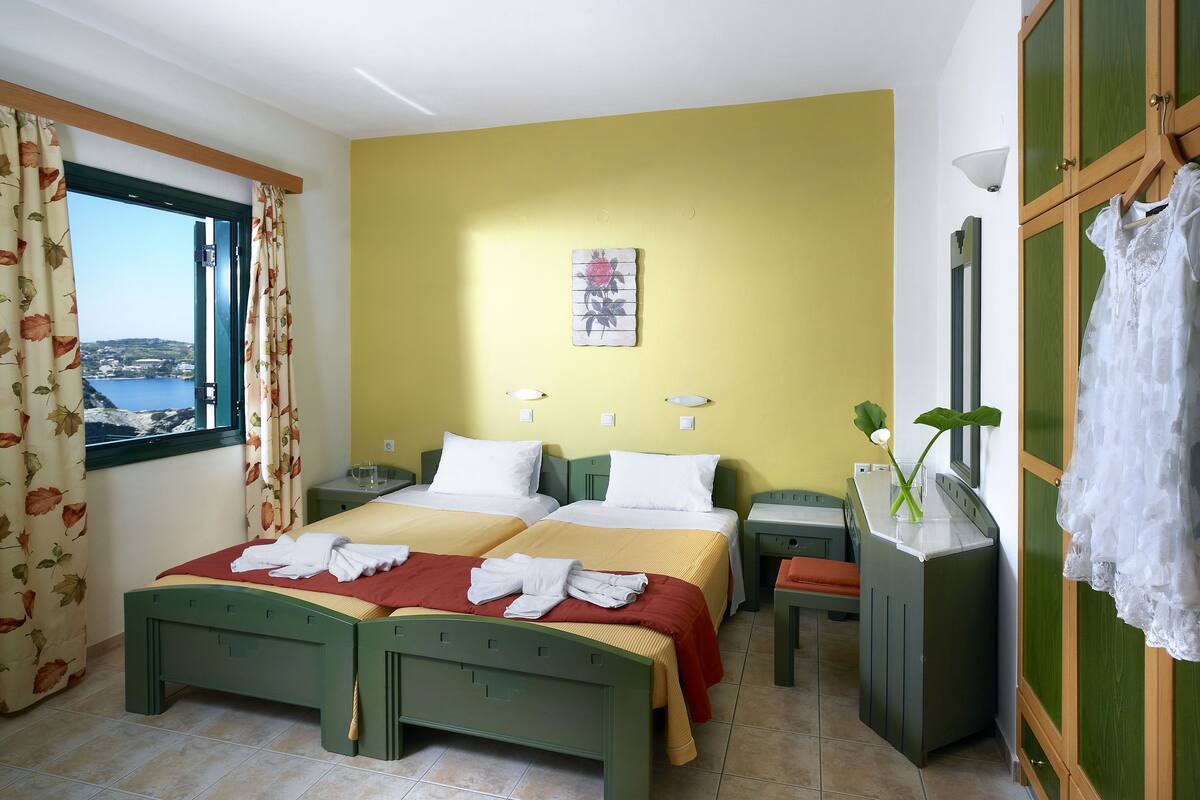 Un lit double avec deux oreillers, une table de chevet, un petit bureau avec une chaise, un miroir et une fenêtre avec des rideaux.