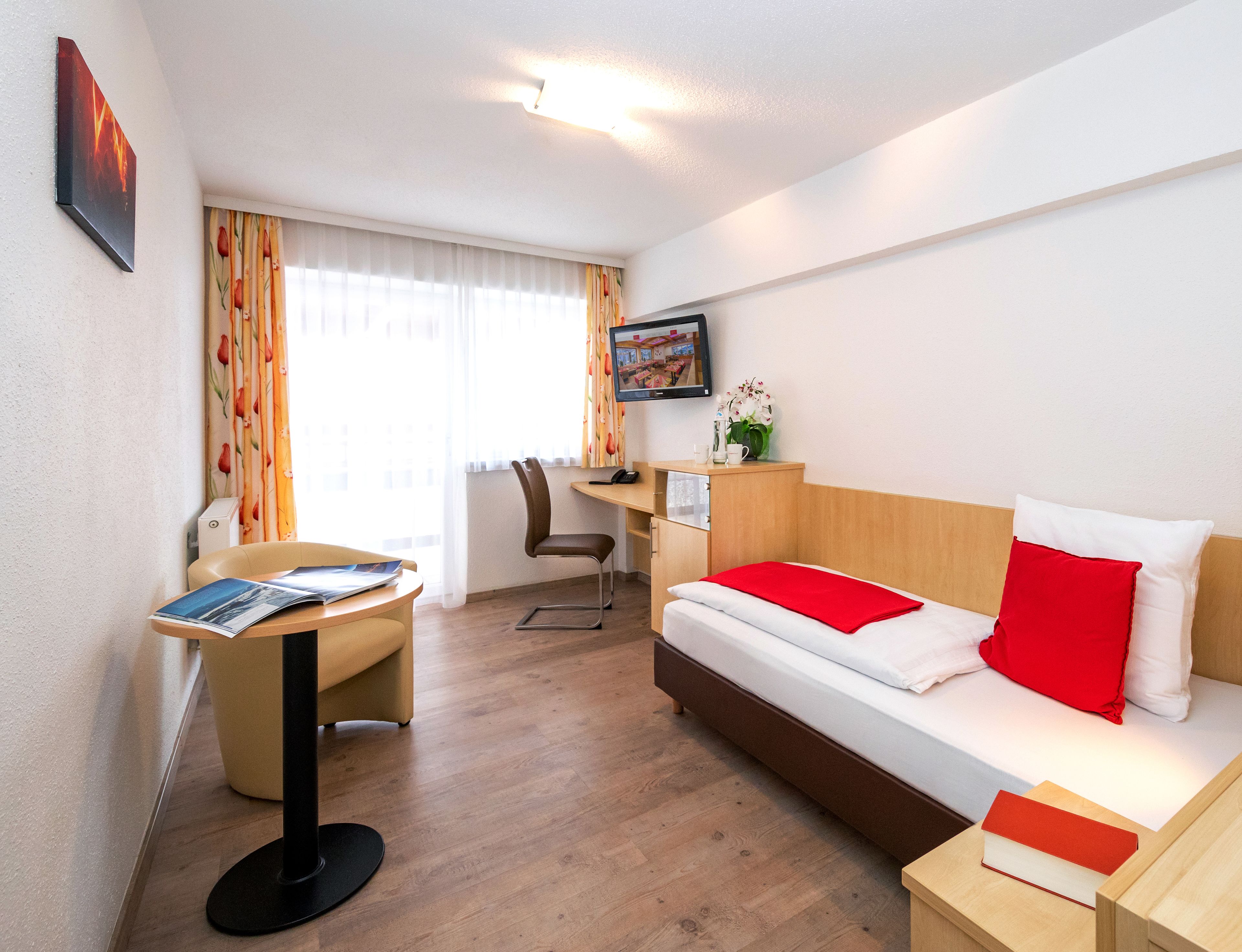 A hotel room with a bed, a desk, a chair, a small table, and a TV on the wall.