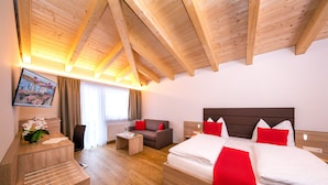 A hotel room with a wooden ceiling, a bed with red pillows, a sofa, a desk with a computer, and a window with curtains.