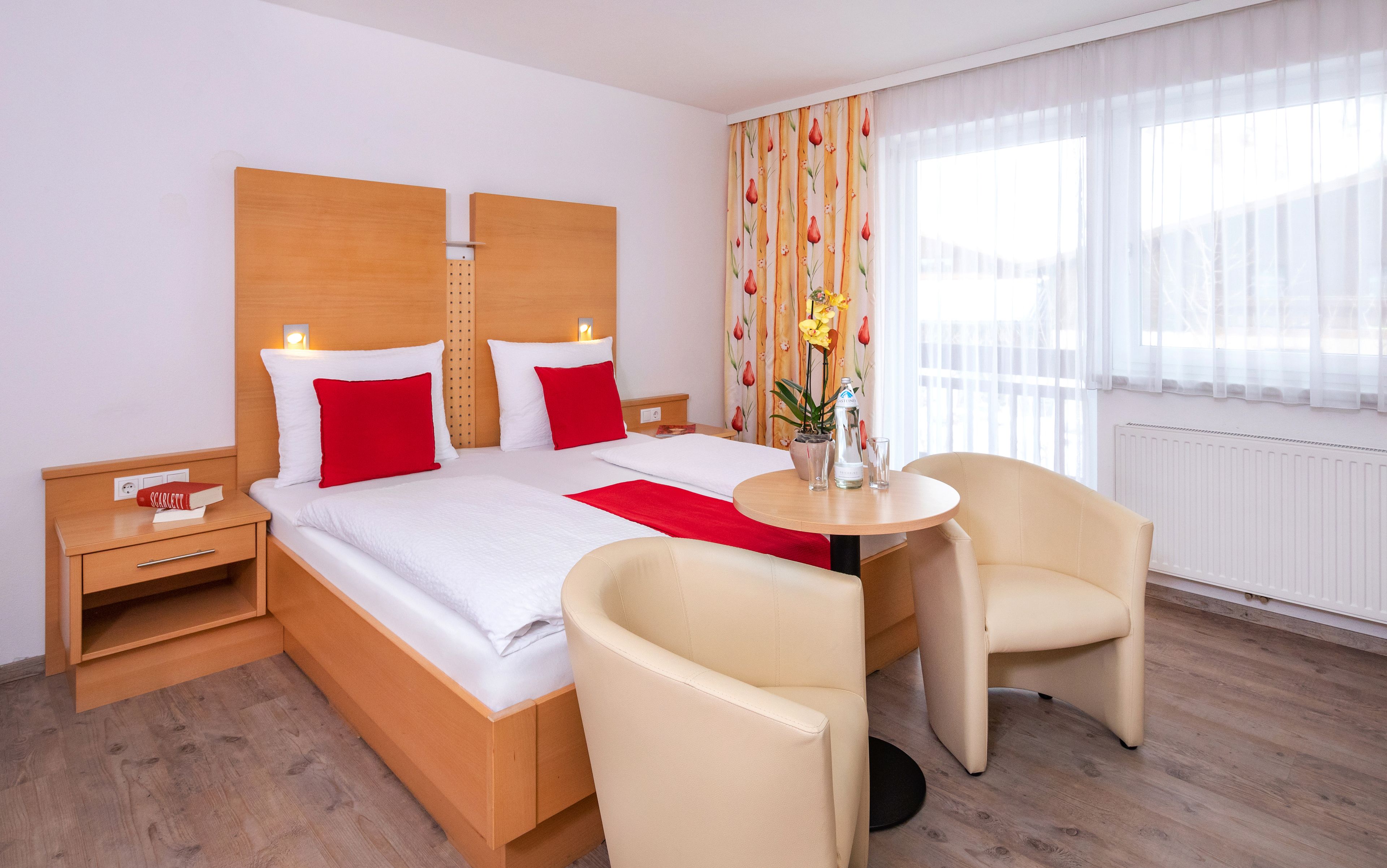 A hotel room with a bed, two nightstands, a bedside table, a chair, a small table, and a window with curtains.