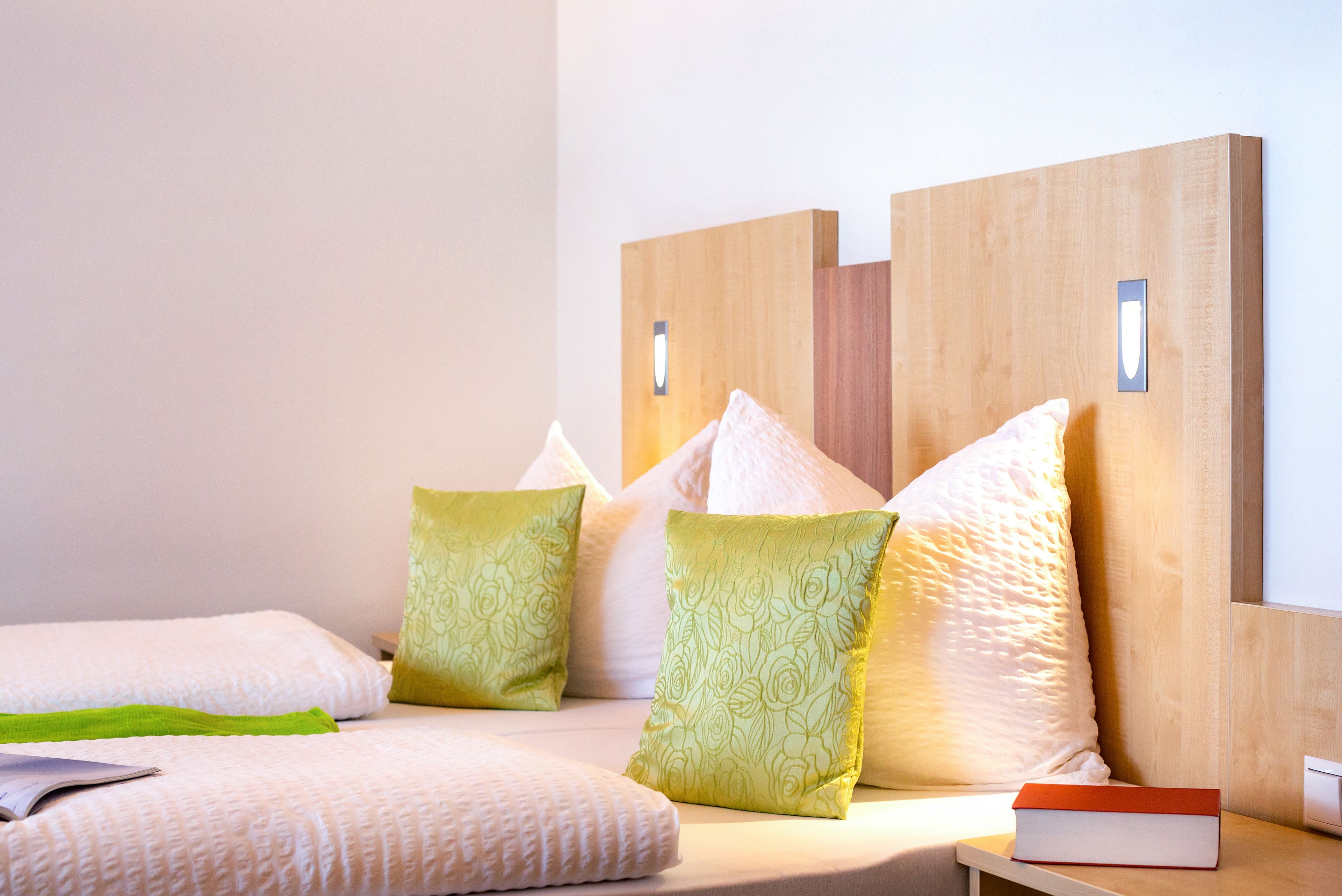 A neatly made bed with pillows, a bedside table with a book, and a wooden headboard with light fixtures.