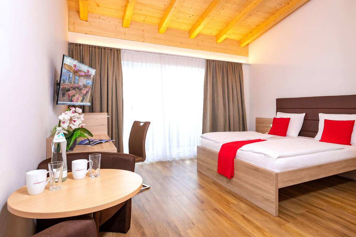 Une chambre d’hôtel avec un plafond en bois, un lit avec une literie blanche et rouge, une table en bois avec deux tasses blanches, un verre et une bouteille d’eau.