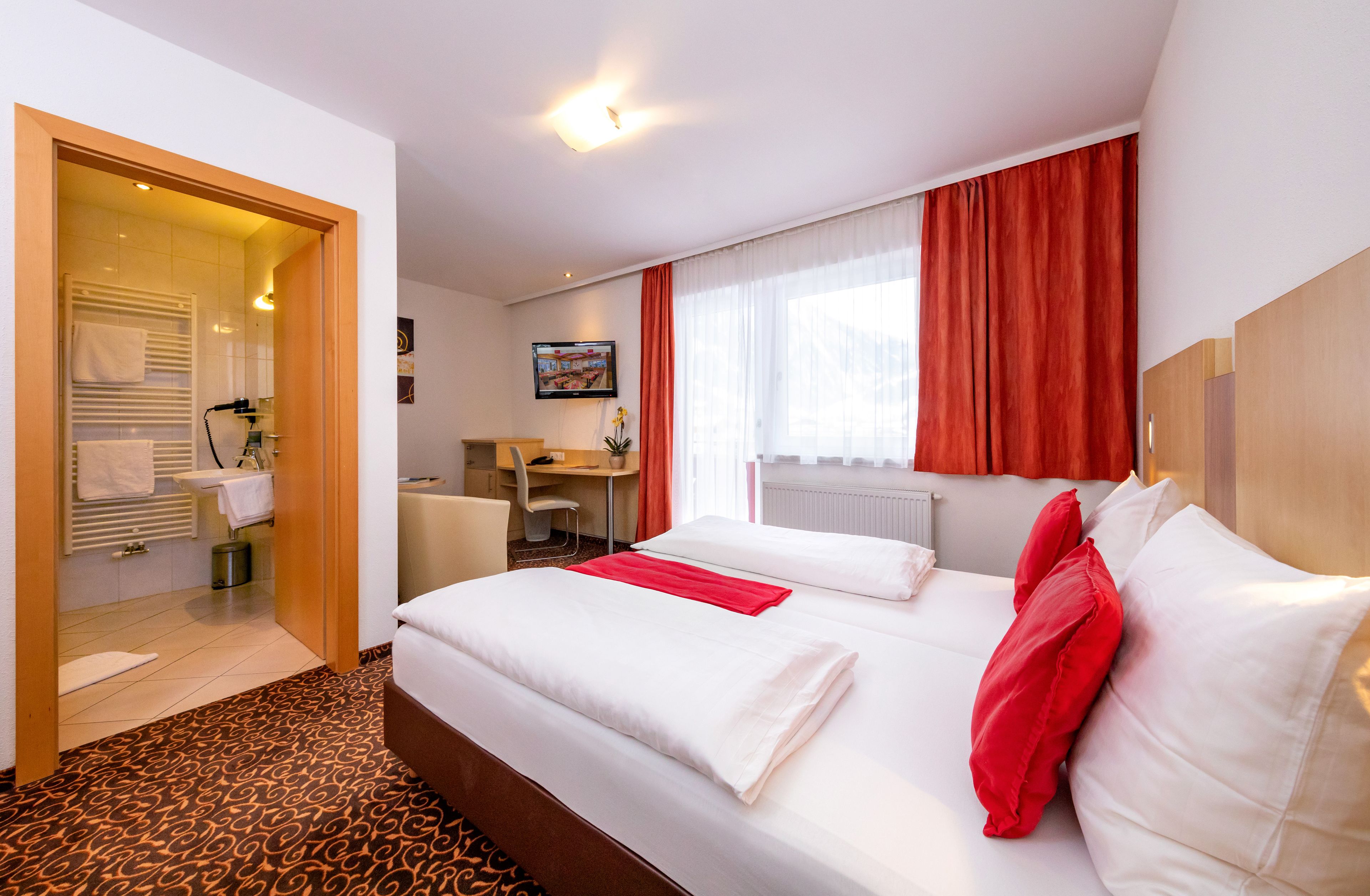 A hotel room with a bed, a desk, a chair, a window with curtains, and a bathroom visible through an open door.
