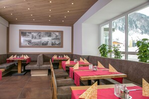 A restaurant with multiple tables and booths, wooden ceiling, and a large window with a view of snow-covered mountains.