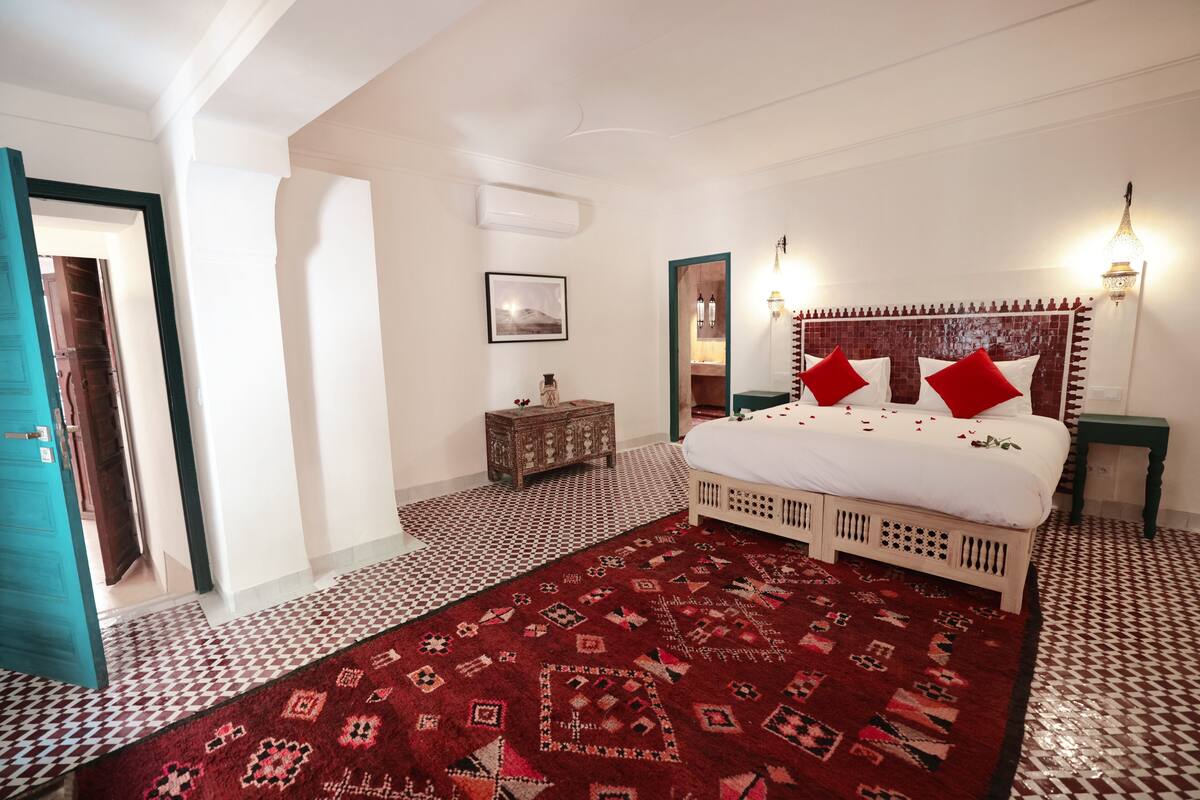A bedroom with a patterned carpet, a bed with red pillows, a nightstand, and a wall-mounted light fixture.