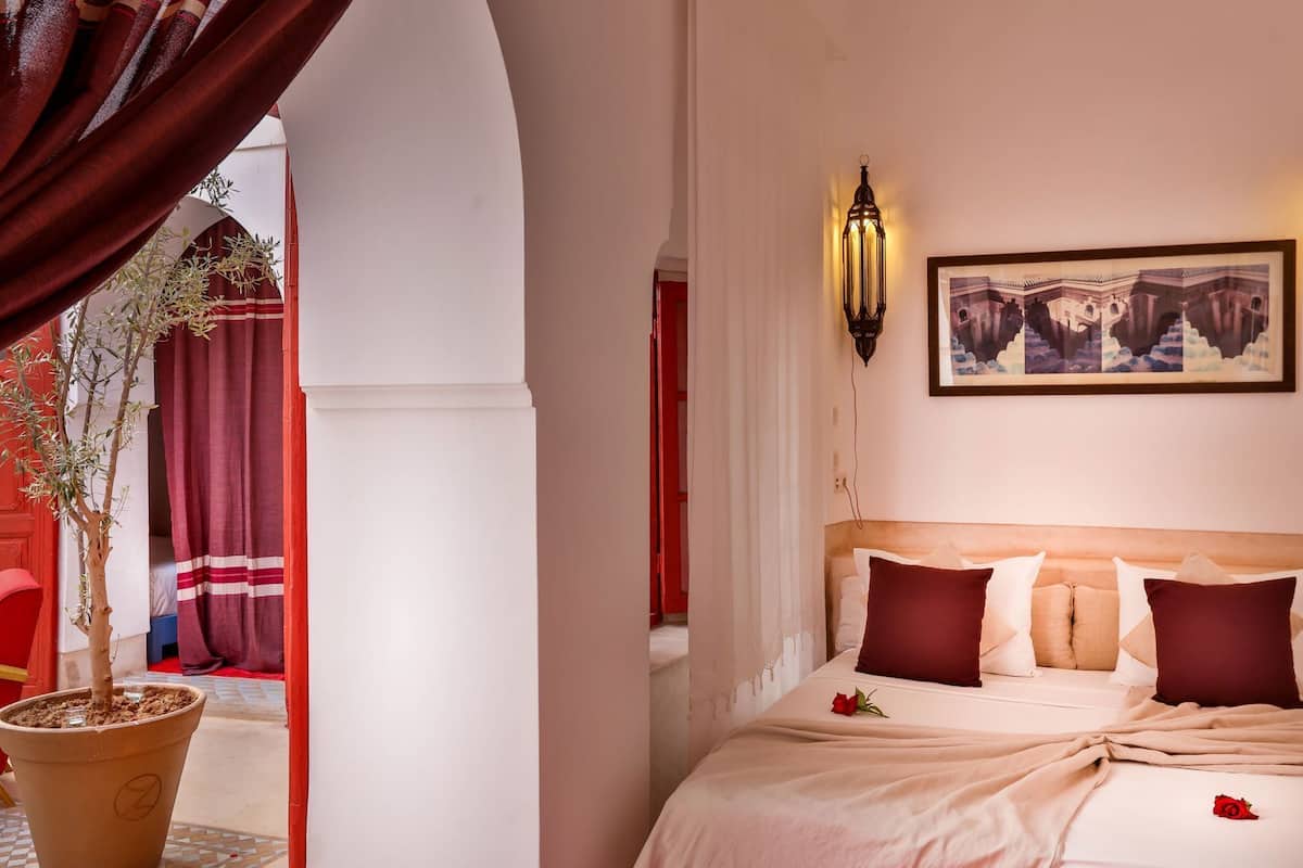 A hotel room with a bed, a potted plant, a wall-mounted light, and a framed picture.