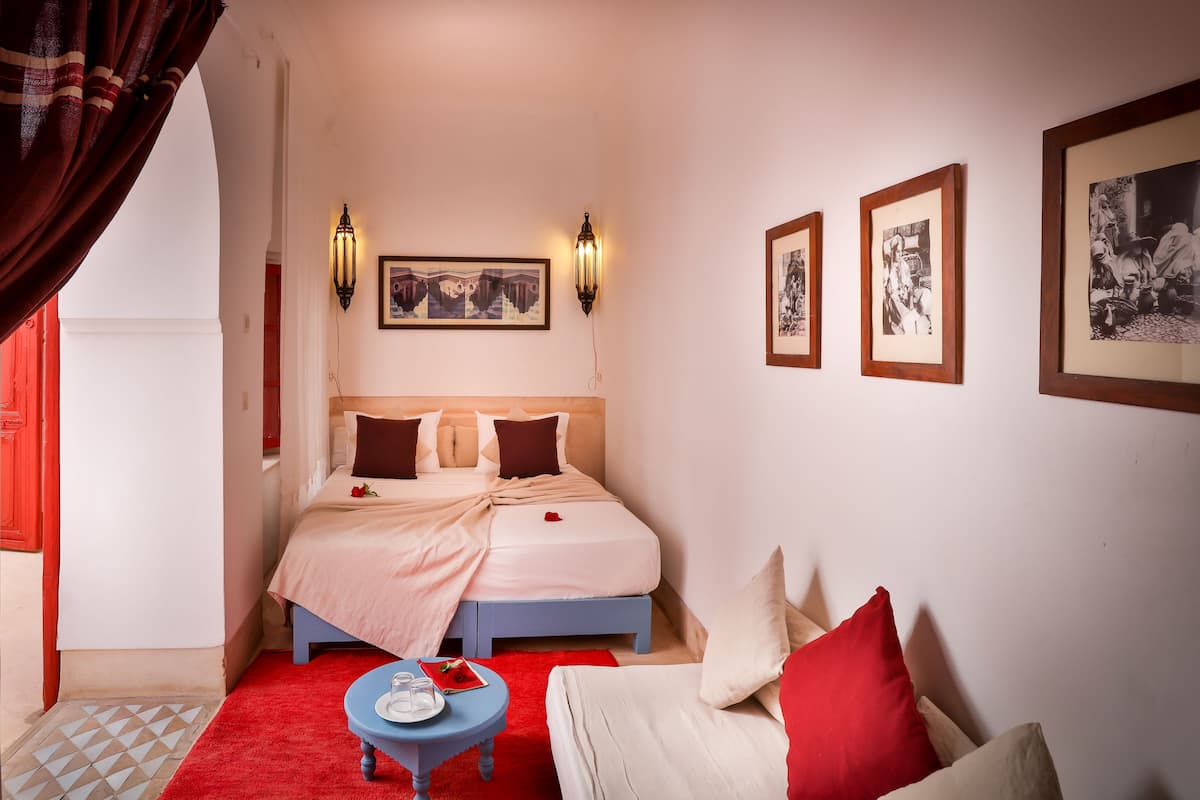 A hotel room with a bed, a small table with a glass, a red rug, and framed pictures on the wall.