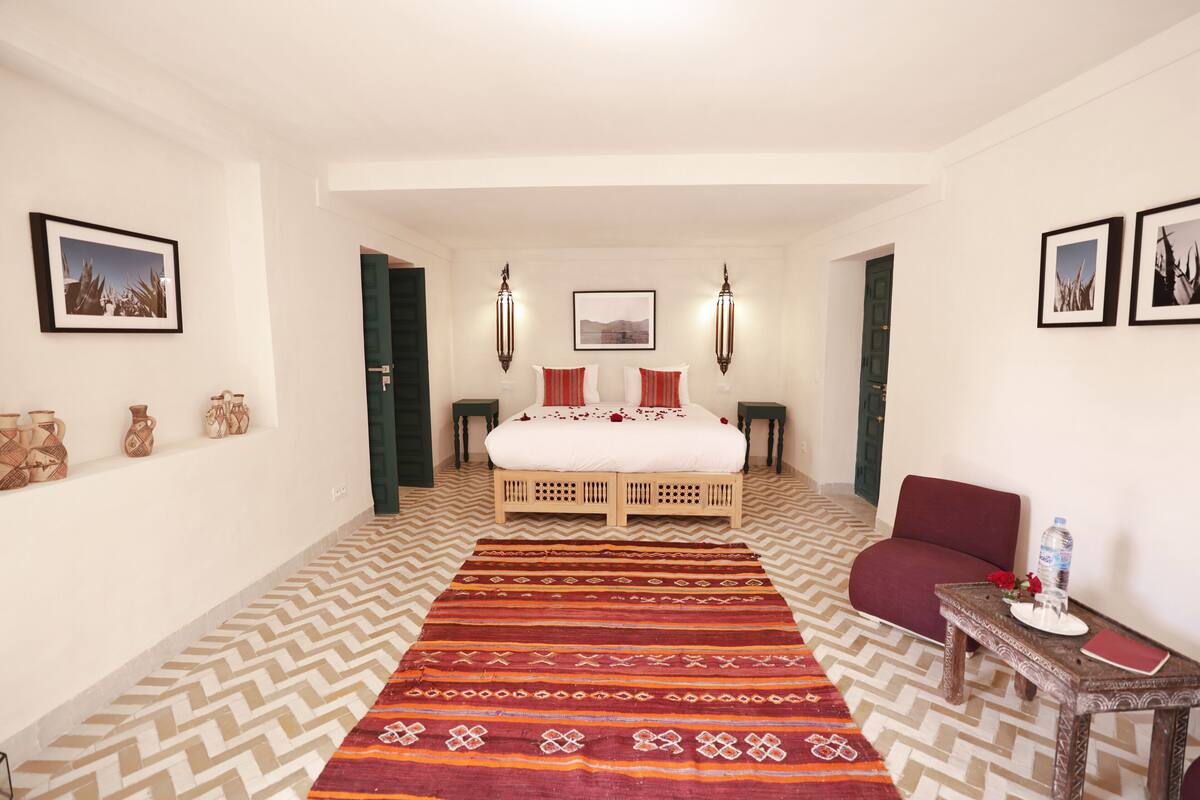 A hotel room with a bed, a patterned rug, a chair, a small table with a water bottle, and framed pictures on the walls.