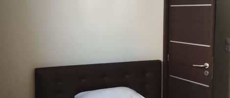 A single bed with a dark headboard, white pillows, and a white bedspread. There is a wall-mounted air conditioning unit above the bed.