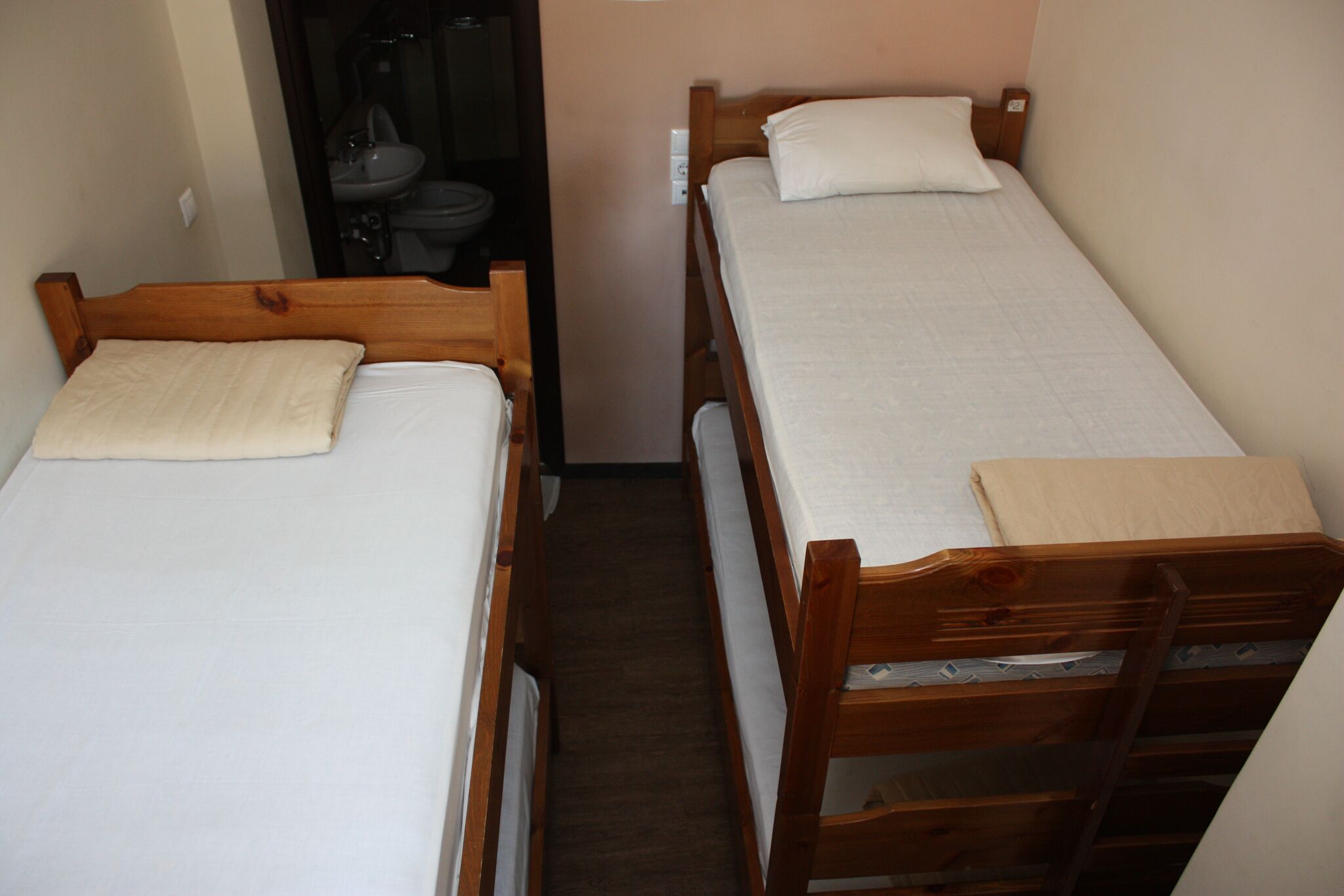 A small, narrow room with two wooden bunk beds, each with a white mattress and a pillow.