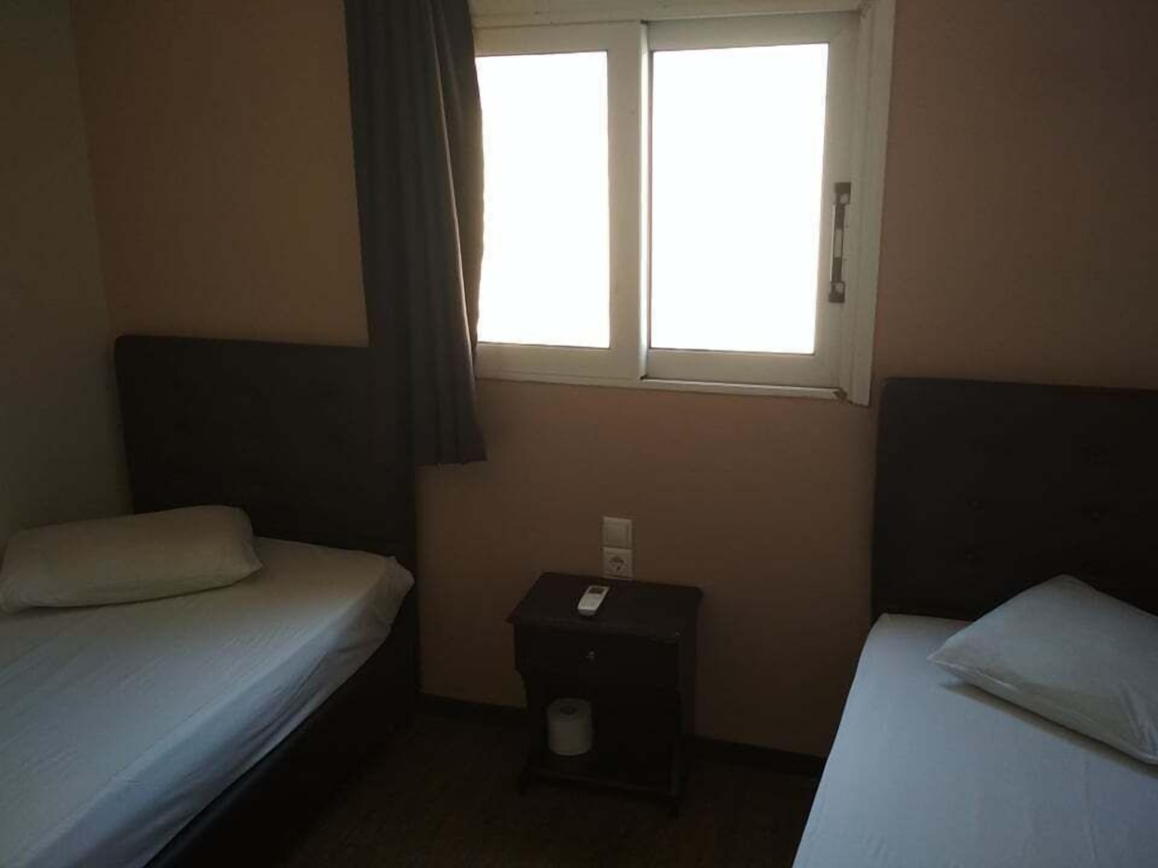 A small, dimly lit hotel room with two single beds, a small table, and a window with curtains.