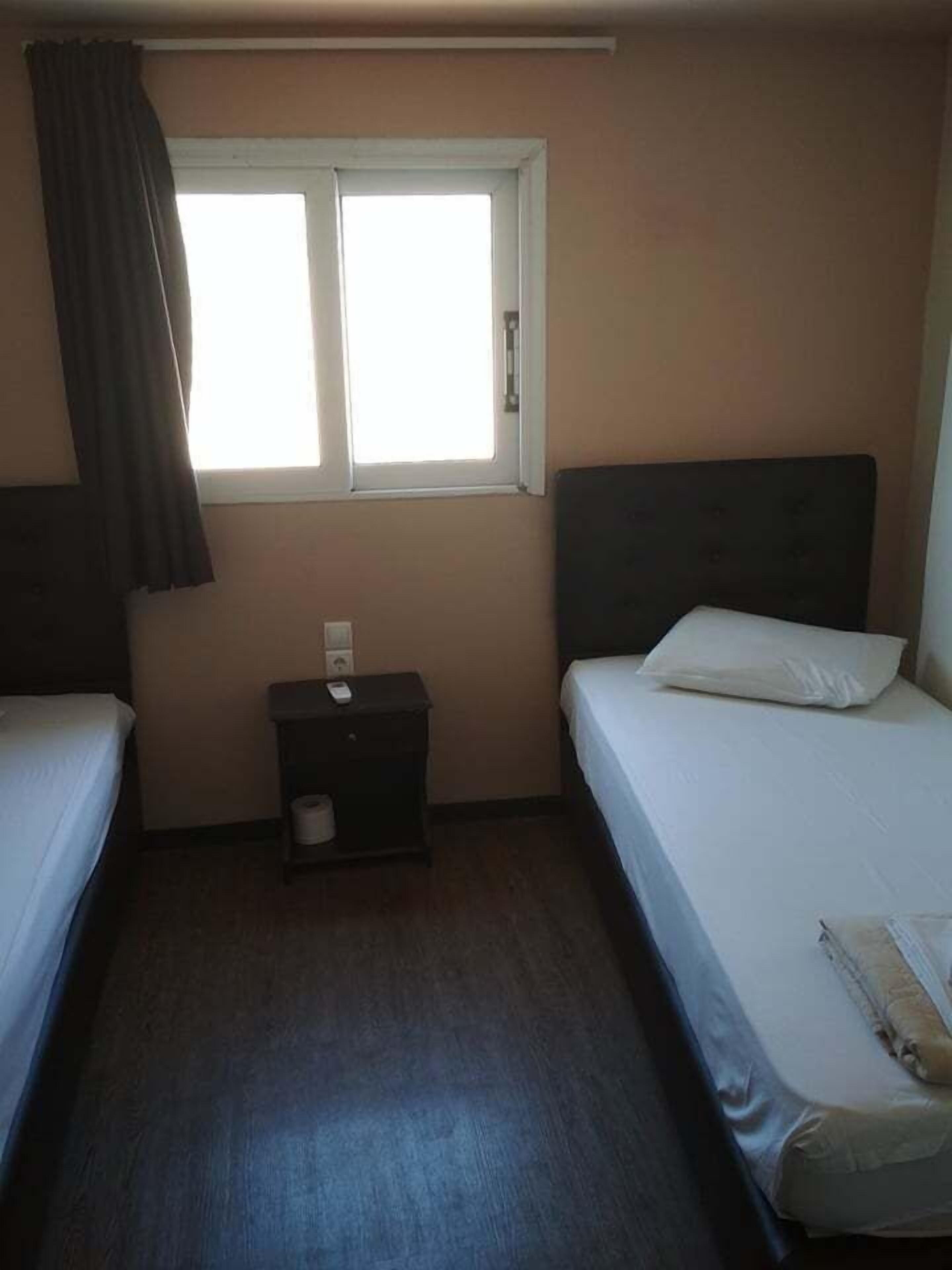 Ett litet, enkelt rum med två enkelsängar, ett litet sängbord, ett fönster med gardiner och en enkel vägg.