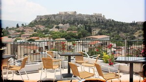 A balcony with wicker chairs and a table, overlooking a historic city with ancient ruins on a hill. - Pella Inn Hostel (Athens)