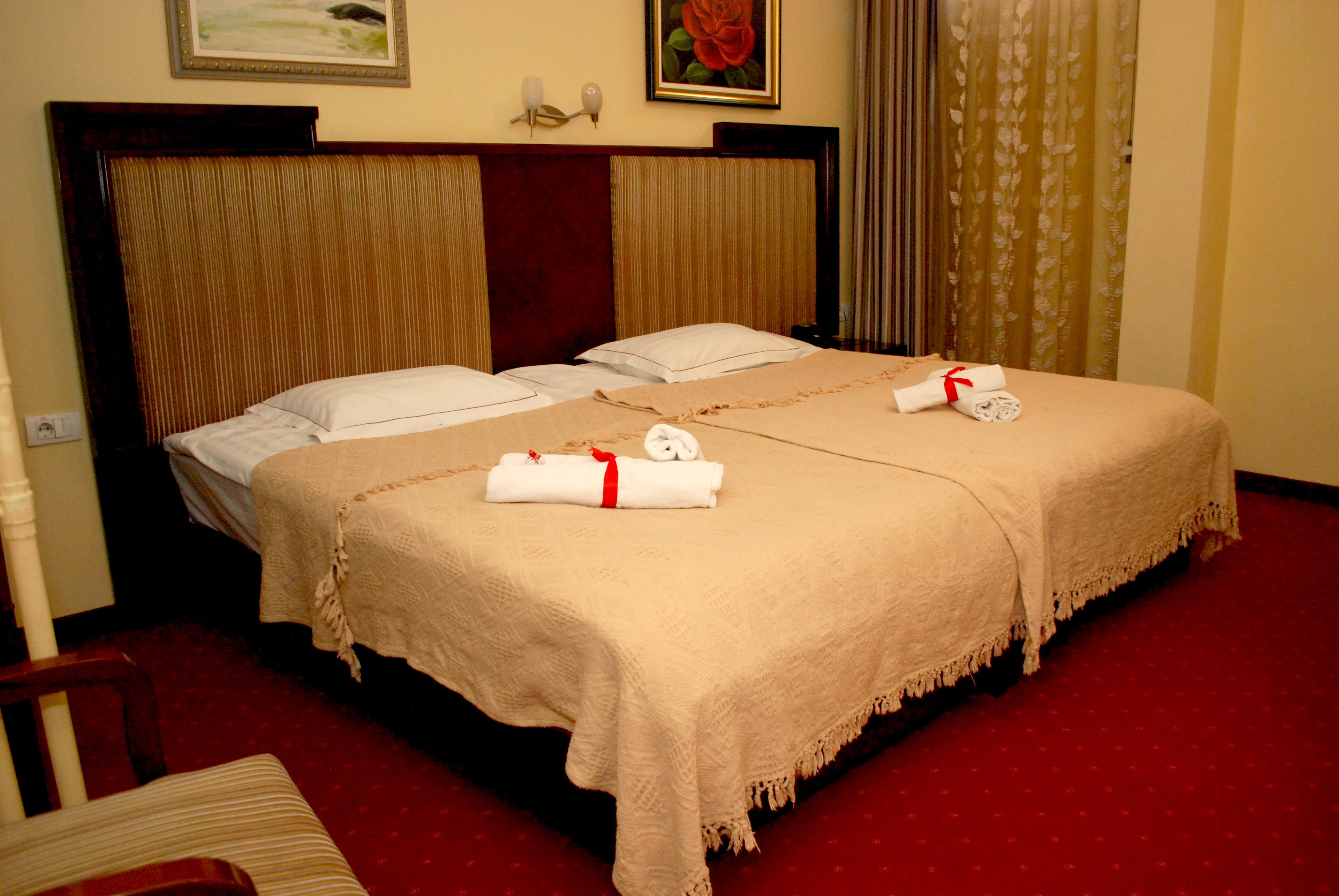 A double bed with two white pillows, a beige blanket, and two rolled towels with red ribbons.