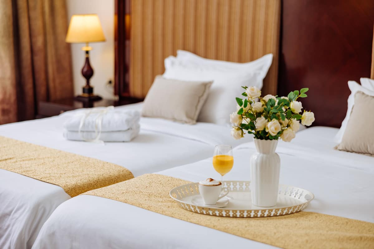 Habitación de hotel con dos camas, un florero con flores, una bandeja con una taza y una copa de vino, y una lámpara al fondo.