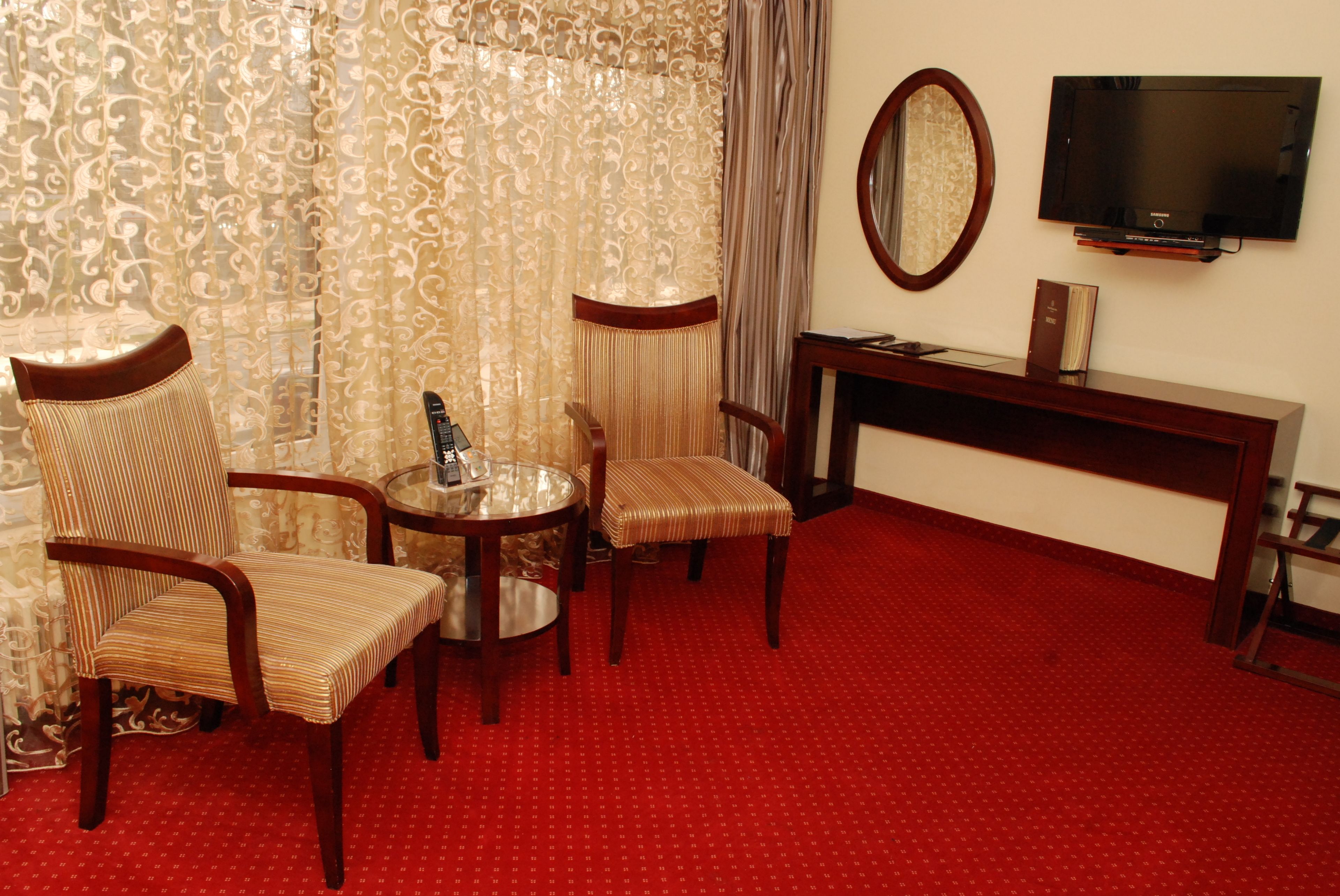 A hotel room with a red carpet, two striped armchairs, a small round table, a TV on the wall, and a wooden console table.