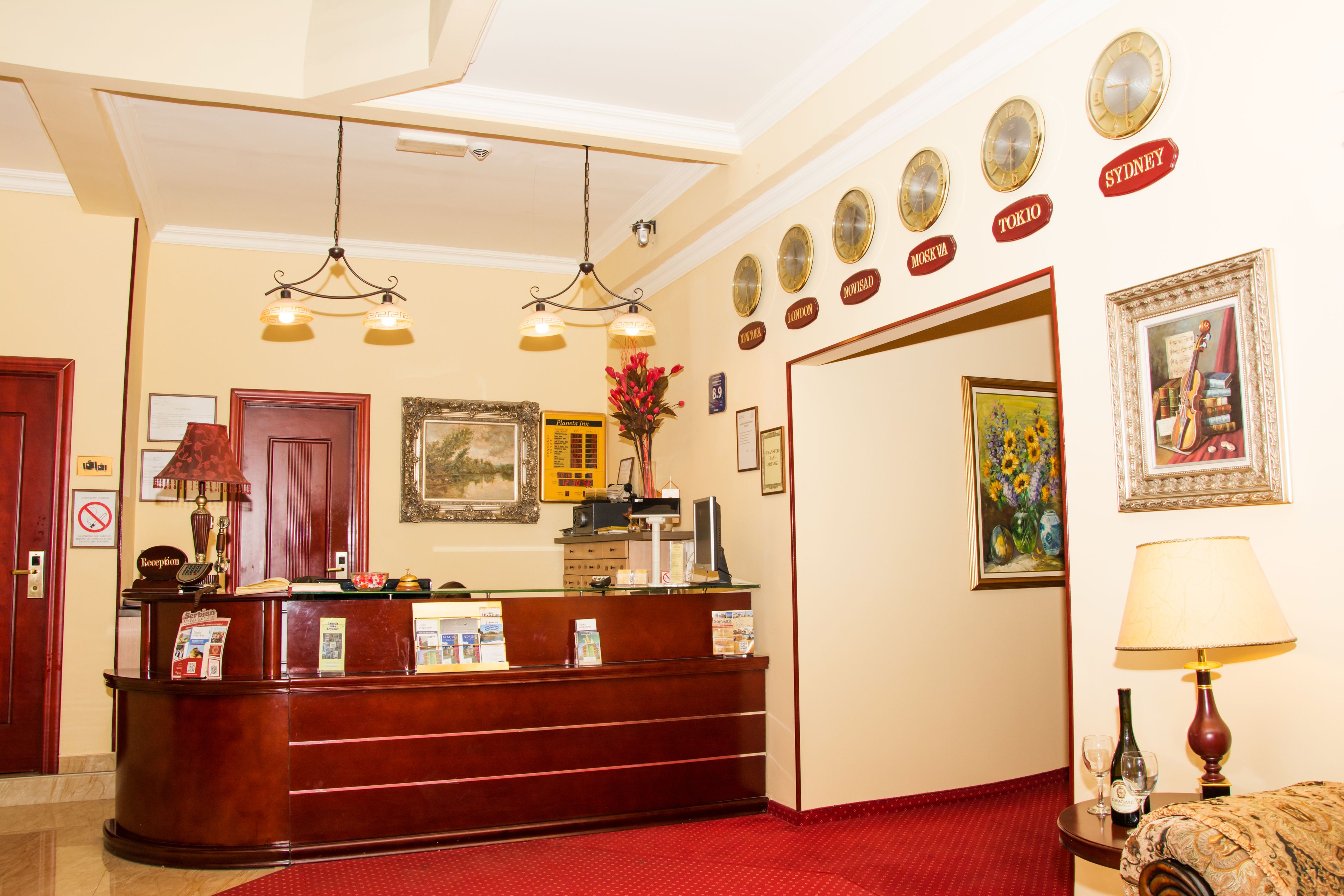 Zona de recepción de un hotel con un mostrador de madera, alfombra roja y decoraciones en la pared, incluyendo relojes y cuadros enmarcados.