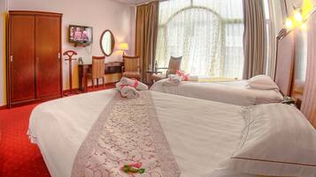 A hotel room with two beds, a desk with a chair, a wardrobe, a television, and a large window with curtains.