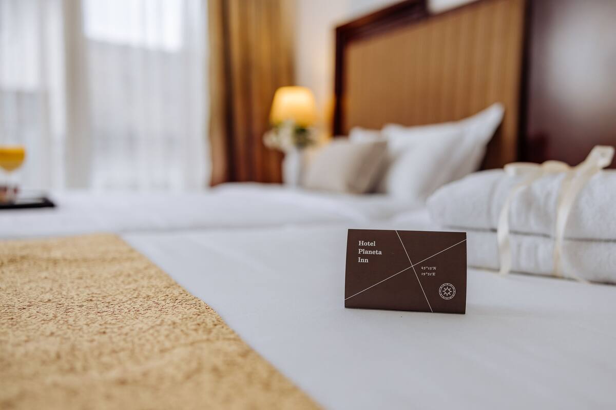 Una cama bien hecha con sábanas blancas, una placa marrón con el nombre del hotel y una lámpara al fondo.