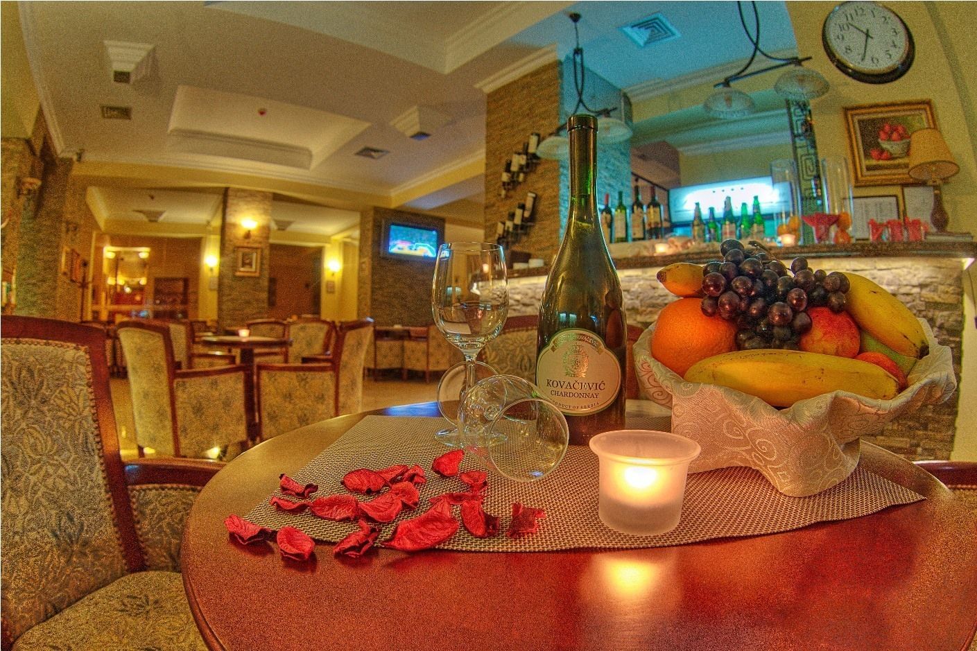A dining table with a bottle of KOVACEVIC Chardonnay, a candle, and a bowl of fruit.