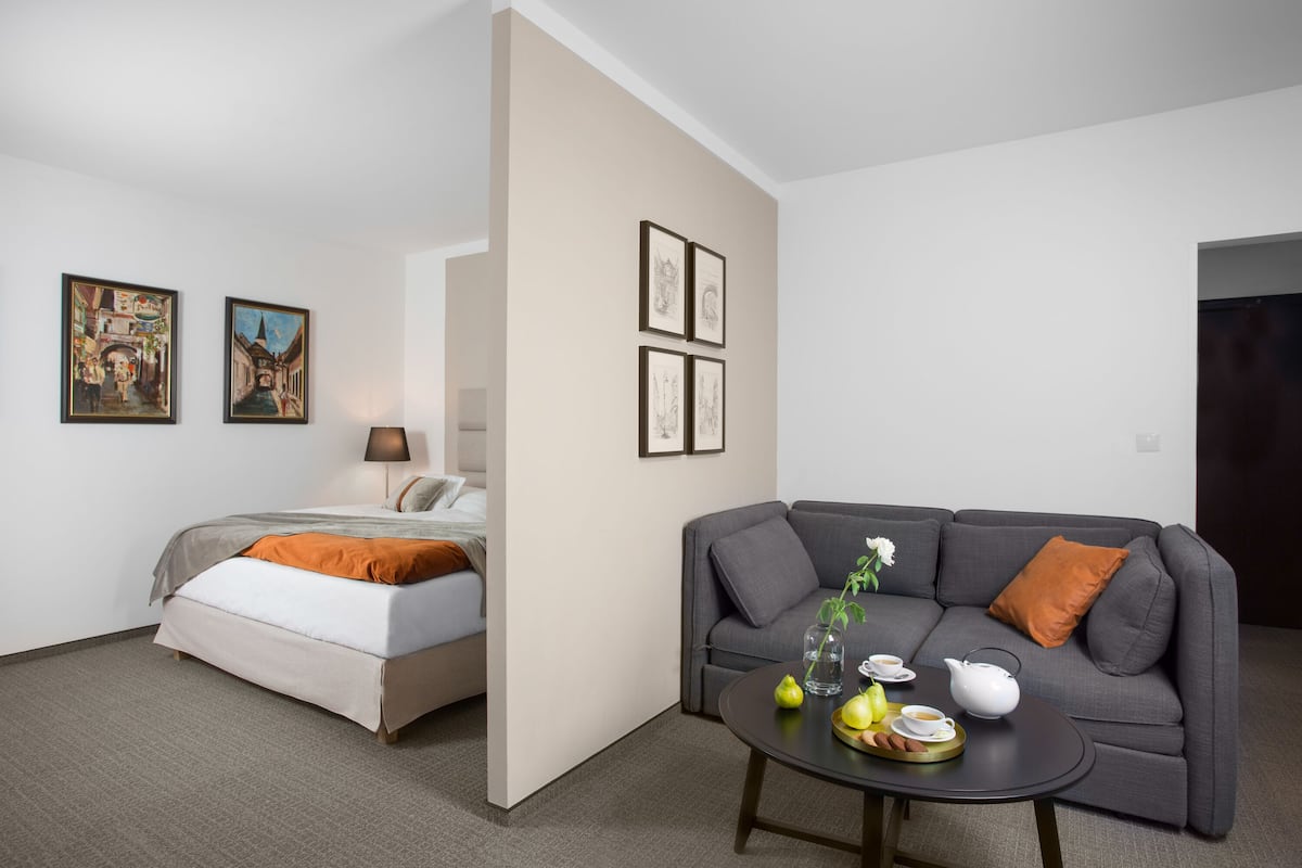 Une chambre d’hôtel moderne avec un lit, un canapé gris, une table basse avec une théière et des fruits, et des œuvres d’art encadrées accrochées aux murs.