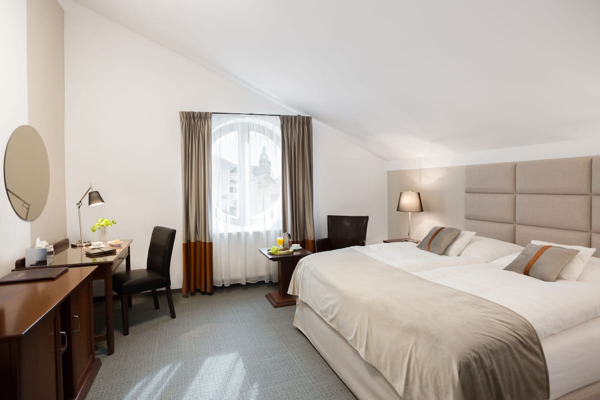 Une chambre d’hôtel avec un grand lit, un bureau avec une chaise, une petite table avec une lampe et une fenêtre avec des rideaux.