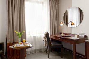A hotel room with a wooden desk, a chair, a bedside table with a tray of breakfast, a lamp, and a large mirror.