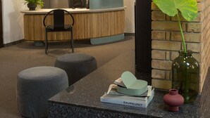 A modern reception area with a wooden desk, a black office chair, and a reception counter. There is a potted plant, a vase, and some books on the counter.