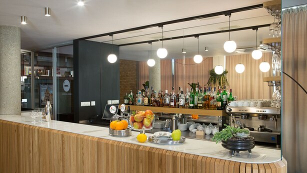 A modern bar area with a wooden counter, various bottles, a coffee machine, and hanging lights.