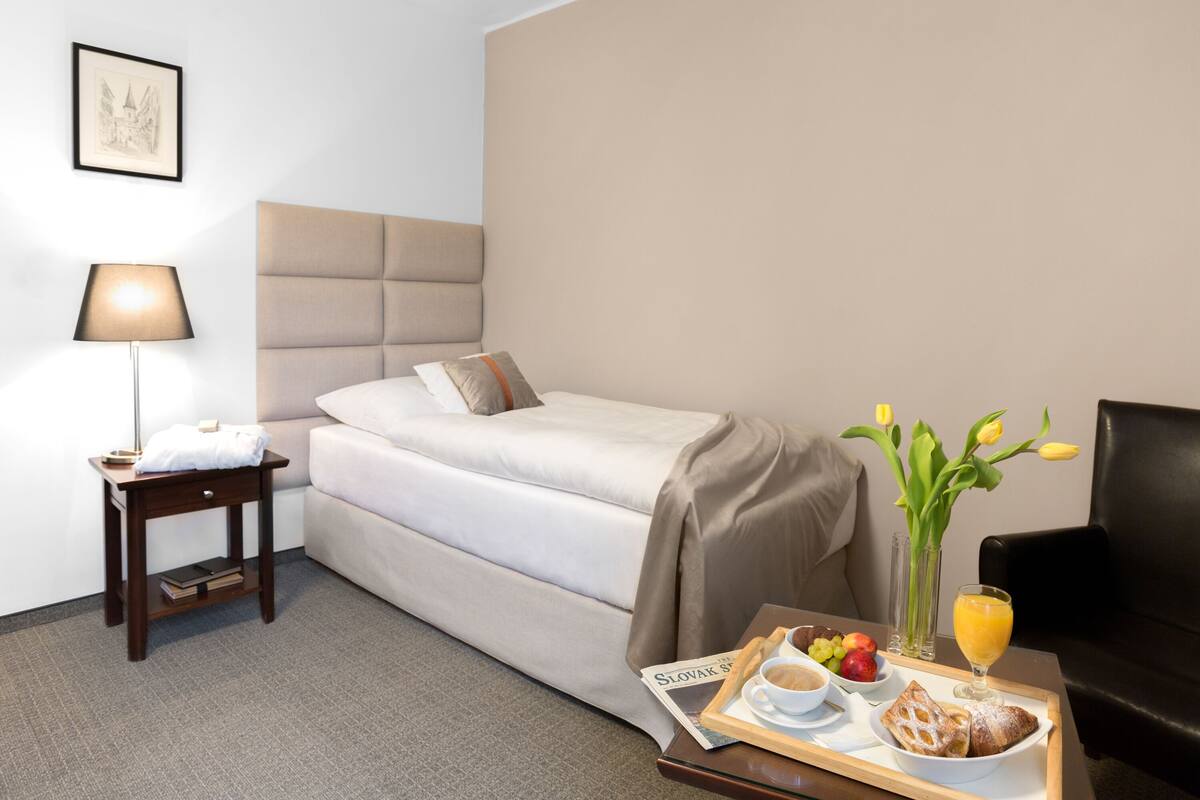 Une chambre d’hôtel avec un lit, une table de chevet avec une lampe, un plateau de petit-déjeuner comprenant des toasts, des fruits et du jus, et un vase avec des fleurs.