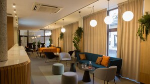 A modern hotel lobby with a reception desk, seating area, and large windows with curtains.