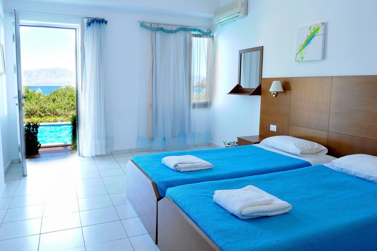Ein Hotelzimmer mit zwei Betten, Blick auf Pool und Berge, sowie ein Badezimmer mit Spiegel und Lampe.