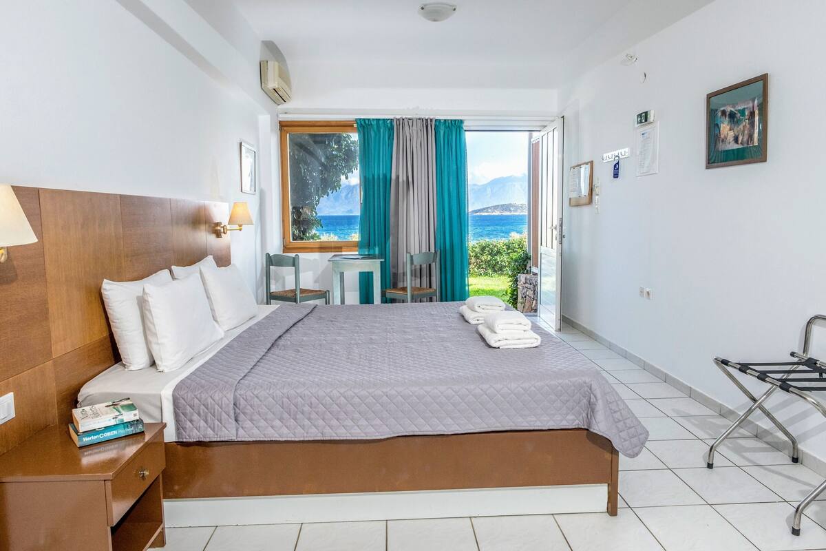 Ein Hotelzimmer mit Bett, Schreibtisch, Stuhl, einem Fenster mit Vorhängen und Blick auf das Meer.