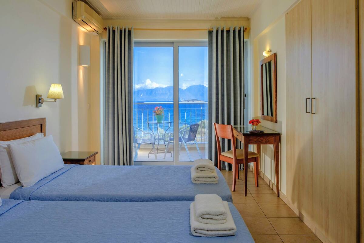 Ein Hotelzimmer mit zwei Betten, einem Schreibtisch, einem Stuhl und einem Balkon mit Blick auf das Meer und die Berge.