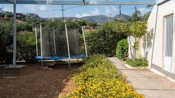 Een overdekt buitengebied met een trampoline, omringd door een tuin vol gele bloemen, en uitzicht op een heuvel.