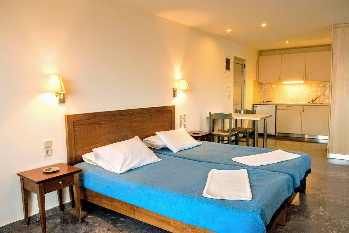 Ein Hotelzimmer mit Doppelbett, Nachttischen, einem kleinen Schreibtisch und einer Küchenzeile im Hintergrund.